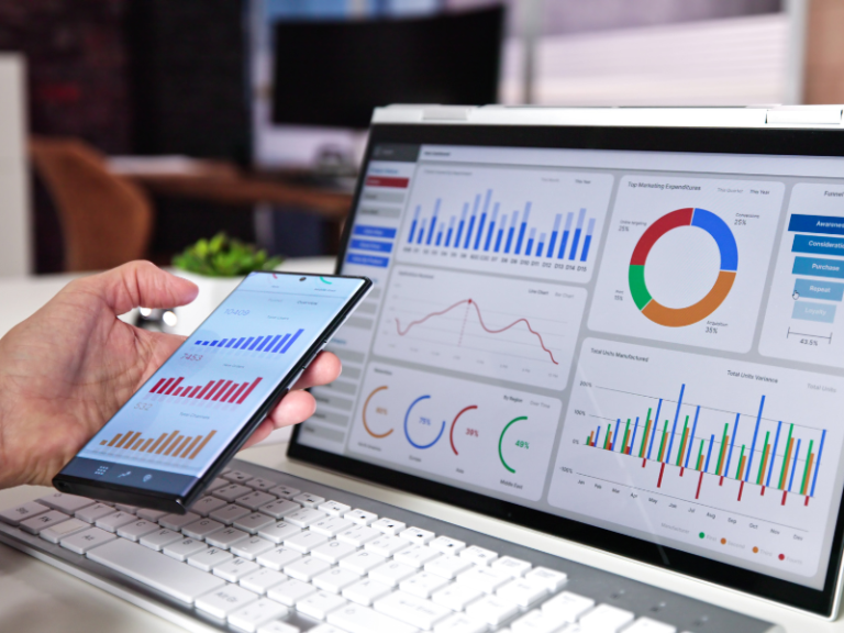 Mano sosteniendo un celular con gráficos de datos, junto a un monitor con un dashboard completo